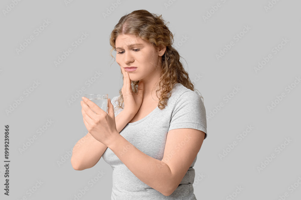 Fototapeta premium Beautiful young woman with glass of water suffering from toothache on grey background