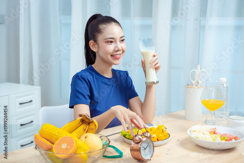 Young woman drinking milk at breakfast table with fruit, cereal, orange juice and clock in bright modern kitchen, healthy morning nutrition and lifestyle concept.