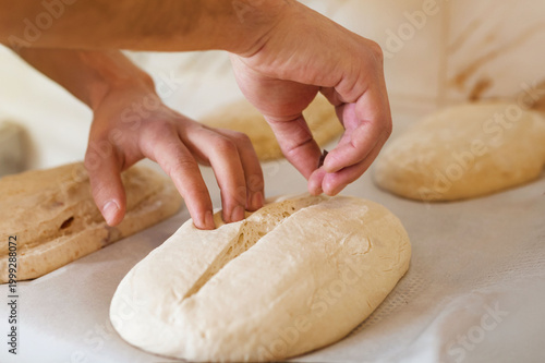Baker scoring raw bread dough with blade, artisan baking process, close up of hands shaping and cutting loaf before baking