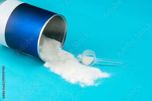 Fine white powder spilled from a blue and white cylindrical container onto a vibrant blue surface. A clear plastic measuring scoop sits within the powder.