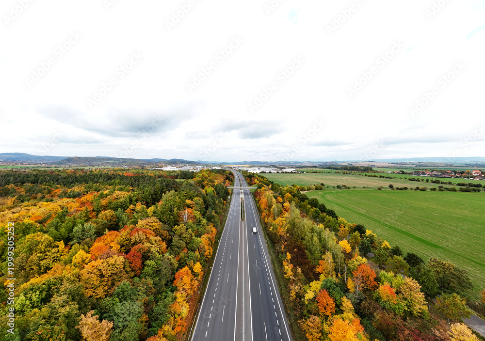Fototapeta premium Wide aerial view of a straight highway passing through a vibrant autumn forest and green agricultural fields under a cloudy sky. Concept of travel and infrastructure.