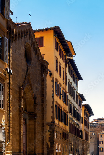 Historic Buildings A Stone Church In The Shadow Next To Yellow And Cream Facades With Closed Shutters In Daylight With A Deep Blue Sky Above