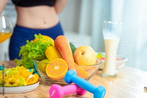 Smiling sporty woman in black sports bra gives thumbs up at kitchen table with fruits, vegetables, juice, water and fitness gear, promoting healthy diet and lifestyle at home.