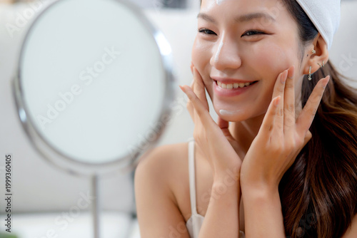 Beauty young asian woman applying cream on facial for wrinkles while looking mirror in living room at home, happy beautiful woman applying lotion moisturizer on cheek for hygiene, skincare and beauty.
