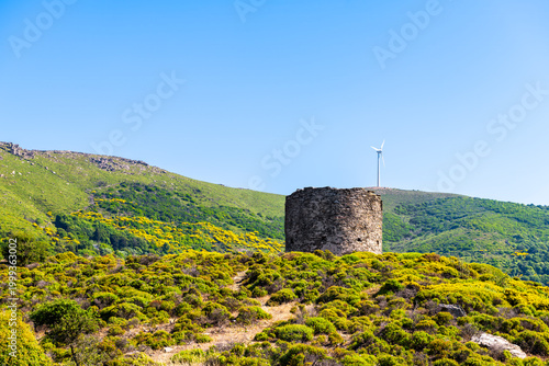Watchtower military tower ruins at Perdiki village on Ikaria island, Greece with windmill turbine for renewable wind energy at longevity blue zone