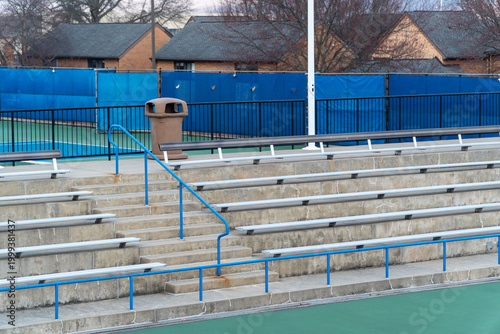 Fotografie Empty stadium bleachers and tennis court with blue fence, trash can, and railings, awaiting spectators for a match or event, showcasing a quiet sports facility