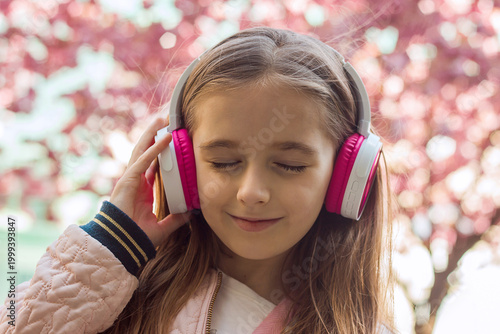 A beautiful little girl listens to music on headphones against the backdrop of flowering trees, close-up portrait