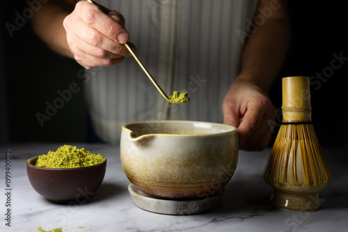 A woman pouring matcha powder into a bowl using a bamboo spoon, preparing traditional Japanese green tea.