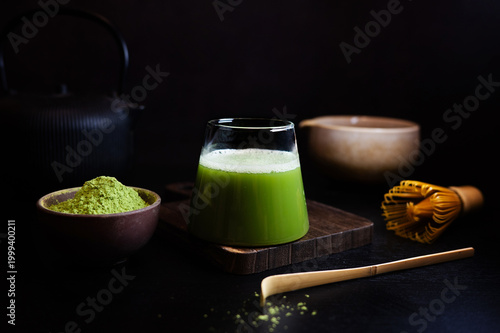Glass of matcha latte with smooth foam next to matcha powder, bowl, bamboo whisk and spoon.