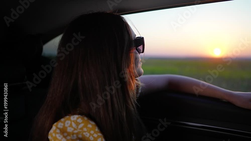 Young woman in sunglasses travels by car at sunset. Happy traveler looks out car window during summer road trip. Girl enjoys sunset travel in passenger seat. Vacation car ride. She smiles.