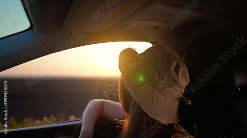Young woman passenger looks out car window at sunset. Girl traveler enjoys summer road trip in vehicle. Female in hat watches golden sun during car ride. Happy woman goes on vacation journey.