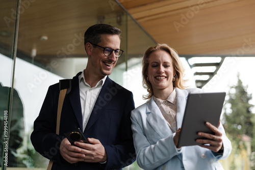 Professionals Smile While Using a Tablet and Smartphone Outside a Modern Building in the Afternoon