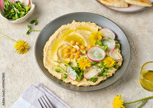 Foto Hummus, an Eastern appetizer made from boiled chickpeas, garnished with microgreens, radish slices, and dandelion flowers, served in a dark ceramic bowl against a light concrete background