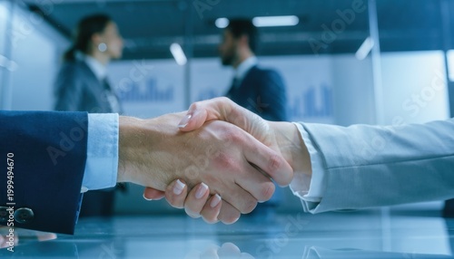 Agreement handshake: Close-up of two business people sealing a deal, their hands clasped in a firm handshake. In the background, other business professionals engaging in a meeting.