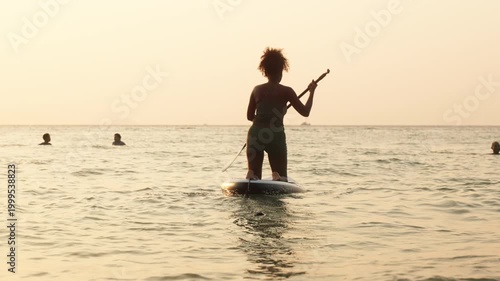 Silhouette of a woman paddleboarding on calm sea during golden sunset, reflecting sunlight on water with distant boat on horizon.