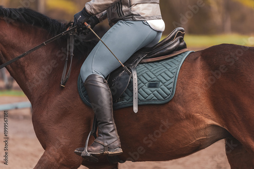 Female equestrian rider on brown horse, close-up of saddle, riding boots and equipment, dressage or show jumping concept. Luxury lifestyle. Premium equipment. Professional posture. Human and animal.