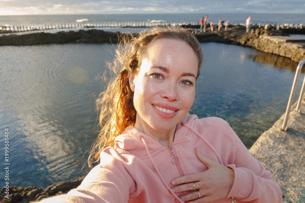 Fototapeta premium A woman smiles as she takes a selfie at sunset by the sea
