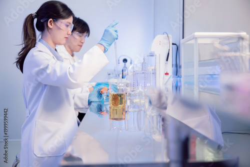 Asian female scientist using pipette to transfer liquid in biotechnology laboratory. Young chemist analyzing liquid chemical reaction in beaker for pharmaceutical industry