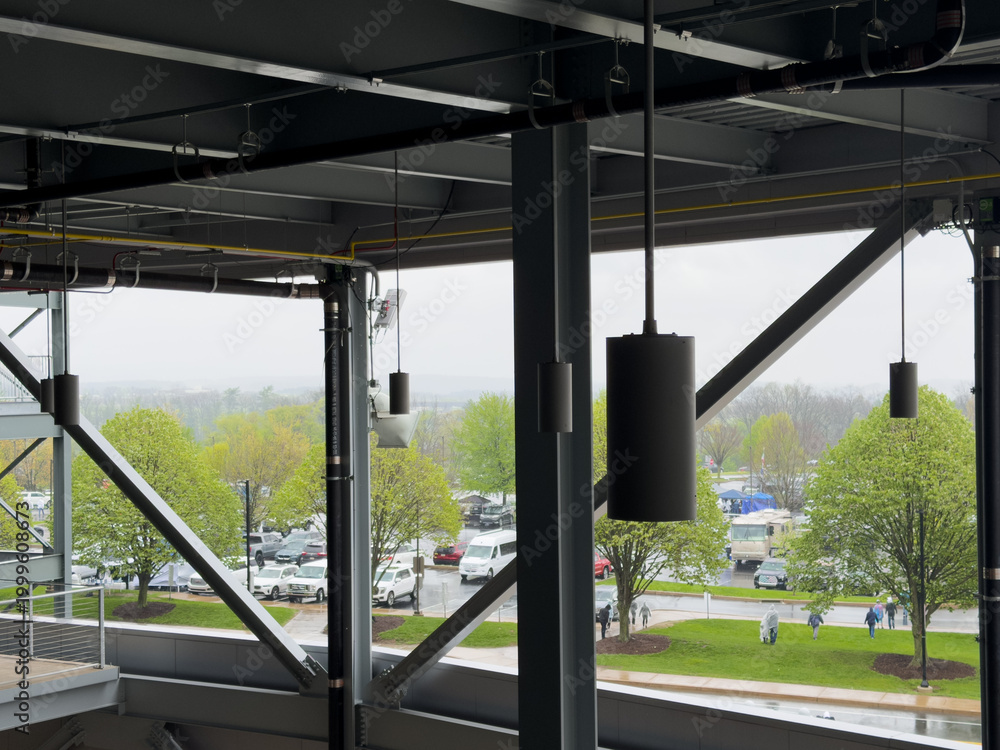 Obraz premium Modern industrial ceiling structure with exposed pipes, speakers, and beams overlooking a rainy parking lot with cars, green trees, and people walking on an overcast day.