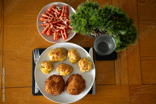 Classic homemade dinner: crispy breaded cutlets, cheesy smashed potatoes and fresh tomato salad on a wooden table. Real home cooking.