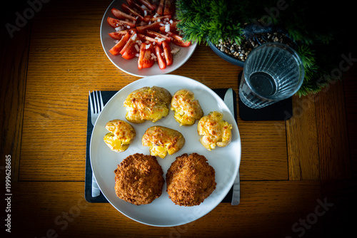 Homemade dinner spread: crispy breaded cutlets with cheesy smashed potatoes, fresh tomato salad and water glass on wooden table. Top view.