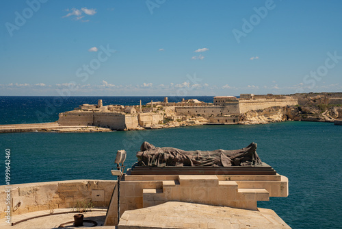 Siege Bell War Memorial with bronze soldier statue in Valletta. View of Grand Harbour and Fort Ricasoli in Malta.
