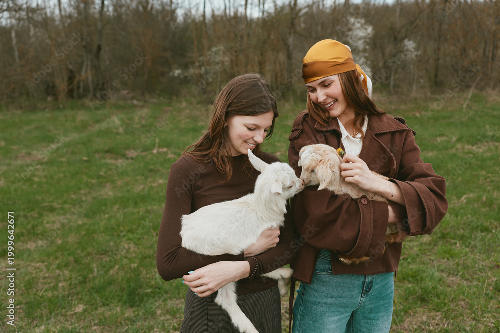 Fototapeta premium Lifestyle scene with two women outdoors holding a kid goat, sharing a gentle moment. Natural light, grassy field, casual wear, connection with animals, promoting wellness and friendship.