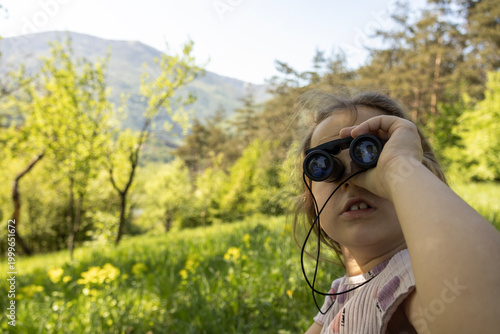 Tableau sur toile Portrait of young girl holding binoculars while exploring sunny countryside meadow surrounded by spring greenery and distant mountains in warm natural daylight