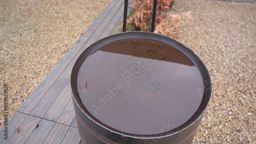 Water-filled barrel top with visible number 1688, surrounded by gravel path and dried plants in a serene outdoor setting