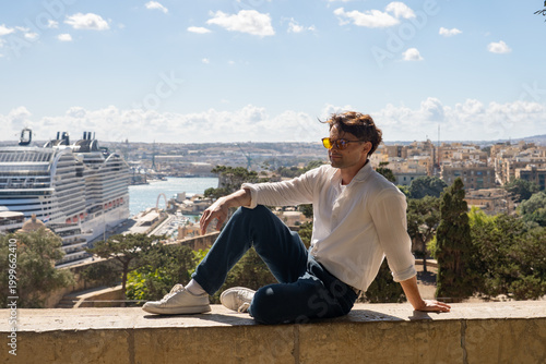 Young man in sunglasses sitting on a historic wall with a giant cruise ship in the background. Mediterranean travel concept in Valletta, Malta.