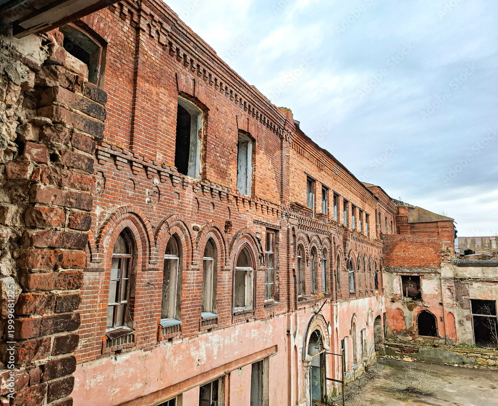 Fototapeta premium Decaying historic industrial building with weathered brick and arched windows under grey sky
