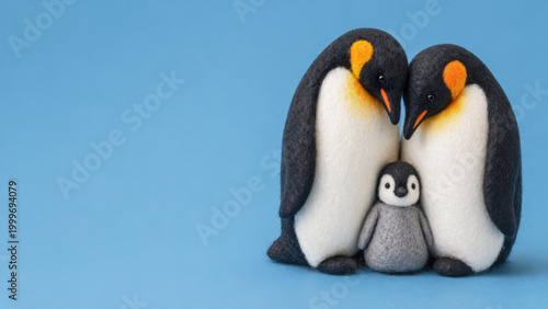 Two handcrafted felt penguins lean together, sheltering a small gray chick between them against a clean blue background, creating a tender, minimalist family portrait.