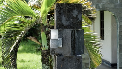 Fotografi A minimalist outdoor wall lamp mounted on a dark textured pillar in a tropical garden setting