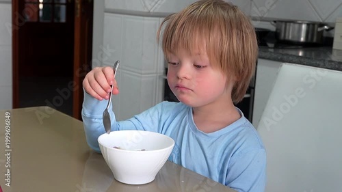 Boy with Down Syndrome in Blue Pajamas Eating Dinosaur Shaped Cereal with Milk in Kitchen