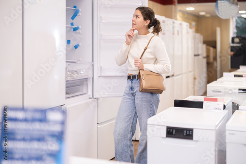 фотографія Young woman buyer examines fridge in detail before buying, checks completeness of household appliances and presence of defects