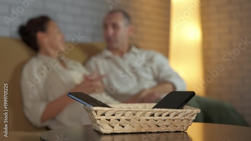 Couple practicing digital detox with phones in a basket. Blurred couple actively communicating on a sofa while their smartphones are put away in a basket in the foreground