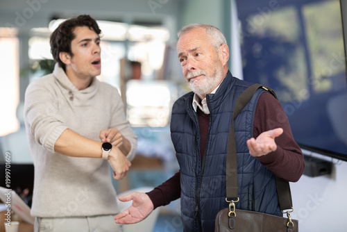 Slika na platnu Young man sternly scolds elderly male colleague for being late for work in offic
