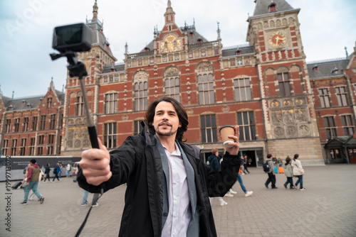 Vlogger recording travel content in amsterdam, holding coffee and selfie stick in front of central station