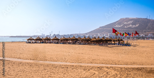 Agadir beach in Morocco, Africa. Sandy beach with straw umbrellas, international flags and sea. Agadir Marina and hill in background. Popular tourist area in Moroccan seaside resort