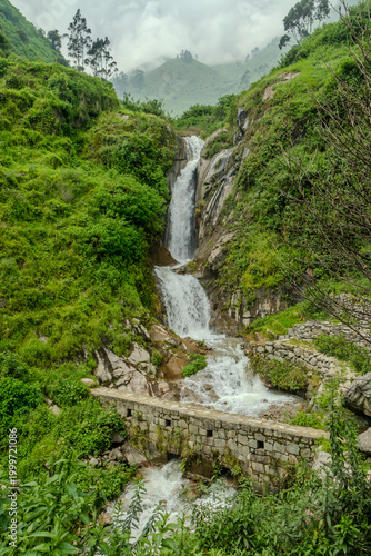 Mountain waterfall flowing over rocks above a small stone bridge in lush green valley