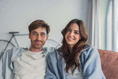 Fényképezés Happy young couple sit close on a comfortable sofa, smiling and relaxed in a bright modern apartment living room, conveying warmth, intimacy and everyday lifestyle moments at home