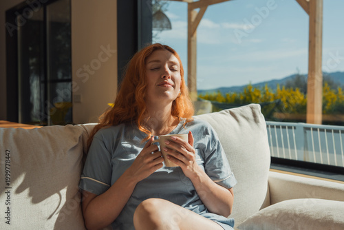 Tableau sur toile A relaxed redhead woman sits on a soft sofa holding a mug, eyes closed and soaking in warm sunlight