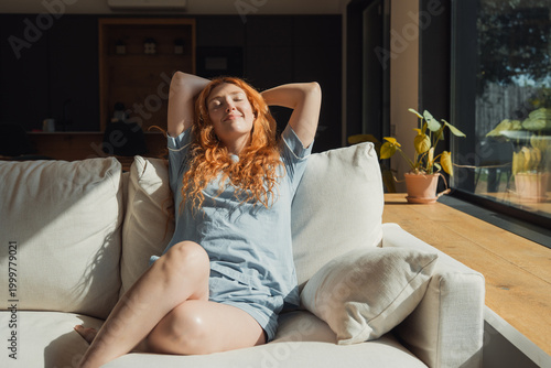 Photos Redheaded woman in casual loungewear lounges on a bright living room sofa, soaking up warm sunlight and enjoying a relaxed, peaceful morning at home surrounded by plants and natural light