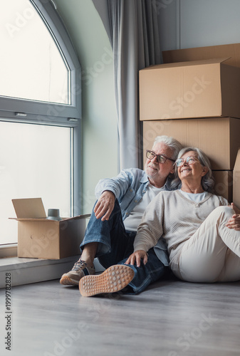 Fotografija A happy senior couple relaxes on the floor beside packed moving boxes, smiling and looking out a window as they settle into their new home during retirement and relocation