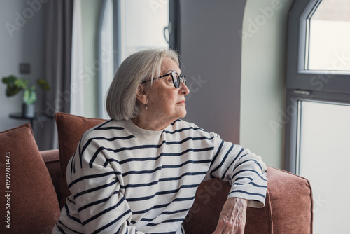 Εκτύπωση καμβά An elderly woman wearing a striped sweater sits on a sofa and gazes out a window, conveying contemplation, quiet solitude and reflective moments in a comfortable home setting with soft natural light