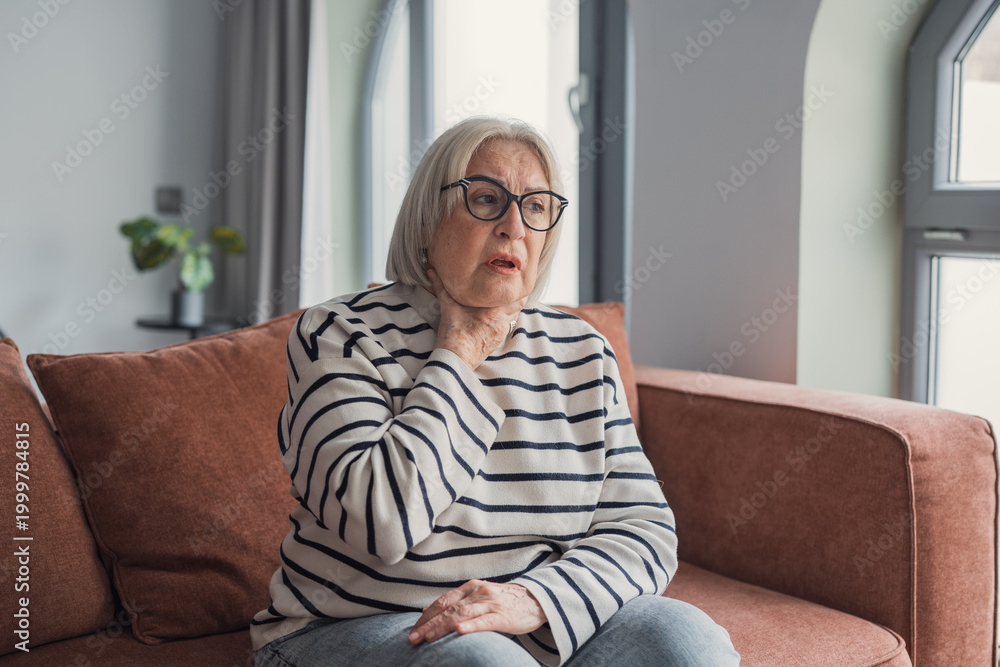 Obraz premium Elderly woman sitting on sofa holding her throat in discomfort at home