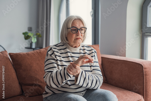 фотографія An elderly woman sits on a sofa at home, holding her wrist with a concerned expression while coping with joint pain and arthritis; a quiet, reflective moment focused on health and aging