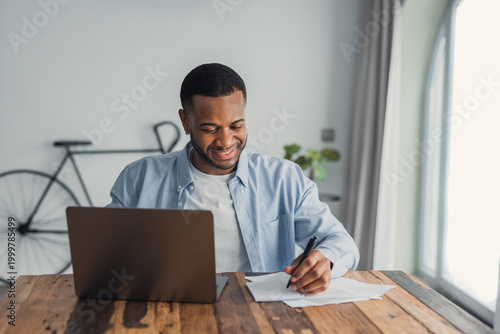 Young man smiling while working from home on a laptop and taking notes on paper at a wooden desk. Casual home office scene with natural light, productivity, focus and remote work atmosphere.