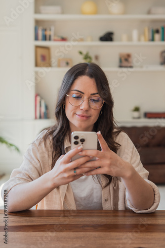Fotografie A young woman sits at a wooden table in a cozy home environment, smiling as she uses her smartphone for messaging, browsing or remote work