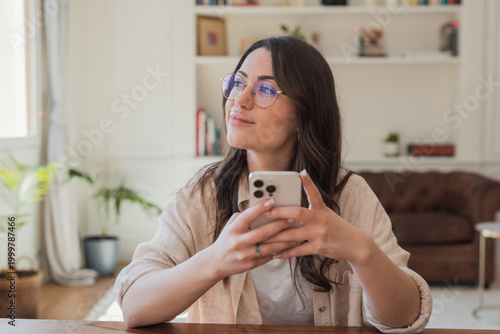 Young woman wearing glasses sits at a table in a bright living room, holding a smartphone and looking thoughtfully out the window, conveying relaxation, contemplation and casual home life.